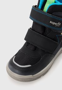 Schwarze Sneaker mit Klettverschluss, strukturiertem Obermaterial, blauen Akzenten, Gummisohle und silberner Zehenkappe. Verfügt über ein atmungsaktives Mesh-Futter.
