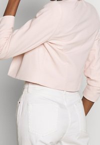Persoon die een lichtroze cropped blazer met gerimpelde mouwen en een hoge taille witte spijkerbroek draagt, gezien van achteren.