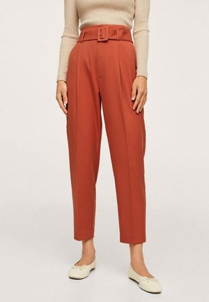 Kobieta ubrana w wysokie, zwężane spodnie w kolorze rdzawym, z dopasowanym paskiem z tego samego materiału oraz beżowy prążkowany długi rękaw.