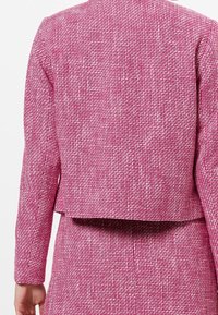 Blazer en tissu tissé rose avec un tissu texturé, un design court et des manches longues, présentant une coupe ample et sans fermetures visibles à l'arrière.