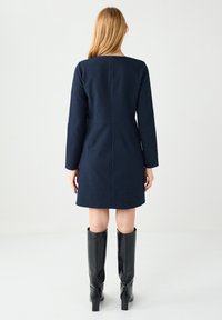 Robe bleu marine texturée à manches longues et coupe droite. Portée avec des bottes noires montantes, affichant un design minimaliste.
