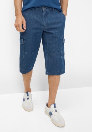 Mand iført blå knælange cargobukser og hvide sneakers med blå accenter, stående mod en hvid baggrund.