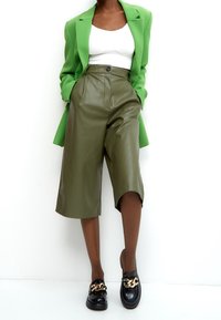 Personne portant un blazer vert vif, un crop top blanc, un short en cuir vert olive longueur genou et des mocassins noirs avec des détails de chaîne dorée.