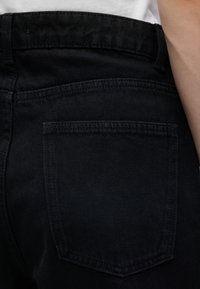 Gros plan sur une personne portant un jean en denim noir montrant la poche arrière et une partie du côté de son bras et de sa taille.
