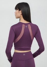 Maglia sportiva a maniche lunghe di colore viola con design a rete sulla schiena, silhouette aderente e dettaglio del logo circolare sul collo, texture morbida.