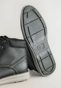 Botas de tobillo de cuero negro con una superficie texturizada, que cuentan con una suela de goma con estrías y costuras blancas a lo largo del borde de la suela.