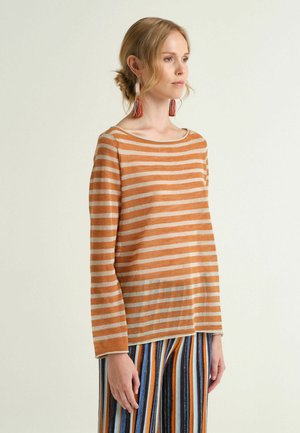 Woman wearing orange and beige striped long-sleeve top and multicolored vertical striped pants, facing slightly right against plain background.