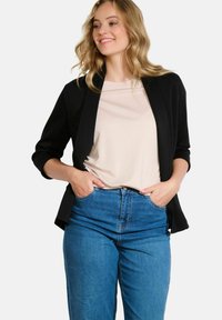 Žena s blond vlasy, oblečená v černém saku, béžovém topu a modrých džínách, s rukama v kapsách, usmívá se a dívá se do strany.