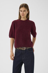 Femme portant un pull en laine bouclée bordeaux à manches courtes, un jean en denim foncé et une ceinture marron sur un fond clair et uni.