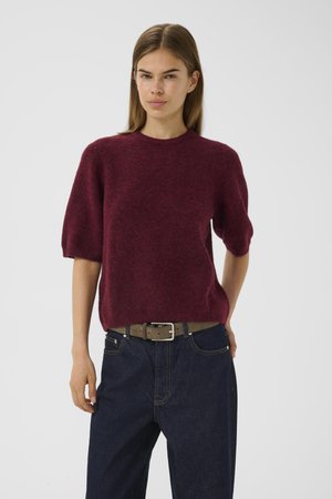 Woman wearing a burgundy short-sleeve fuzzy sweater, dark denim jeans, and a brown belt against a plain light background.