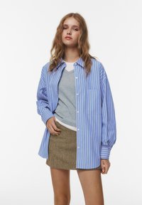 Tânără cu păr ondulat, îmbrăcată cu o bluză albastră în dungi, un pulover gri cu guler în formă de V, un tricou alb pe dedesubt și o fustă mini cu model maro, stă în fața unui fundal alb.