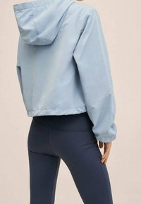 Kvinna i ljusblå huva jacka och mörknavy tajta leggings, sedd bakifrån mot en neutral bakgrund.