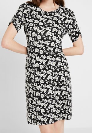 Czarna sukienka za kolano z krótkimi marszczonymi rękawami, pokryta białym kwiatowym wzorem z żółtymi środkami, noszona przez osobę z rękami na biodrach.