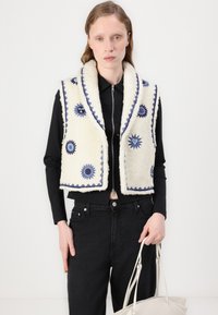 Vit, fluffig väst med blå blommig broderi, scalloped trim och sjalkrage, bärs över en svart topp och matchas med svarta byxor.