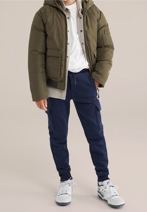 Lapsi, joka pukeutunut oliivinvihreään toppatakkiin, harmaaseen paitaan, valkoiseen t-paitaan, tummansinisiin cargo-housuihin ja valkoisiin lenkkareihin, seisoo kädet osittain taskuissa.