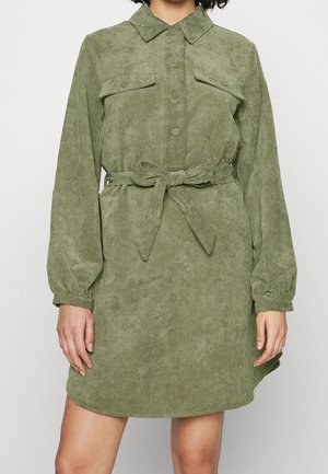 Woman wearing a long-sleeve, olive green corduroy shirt dress buttoned up with a tied fabric belt at the waist.