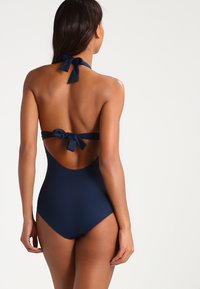 Dunkelblauer Einteiler mit Halterneck-Bändern und offenem Rücken. Glattes Material und hoch ausgeschnittene Beine, die eine elegante Silhouette betonen.