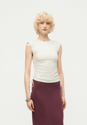 Jeune femme aux cheveux blonds courts et bouclés, portant un haut blanc froncé sans manches et une jupe violette taille haute, debout devant un fond uni.