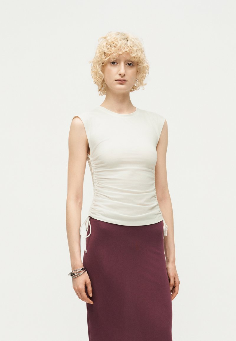 Νεαρή γυναίκα με κοντά κατσαρά ξανθά μαλλιά που φοράει λευκό αμάνικο κορσέ και μωβ φούστα με ψηλή μέση, στέκεται μπροστά σε απλό φόντο.