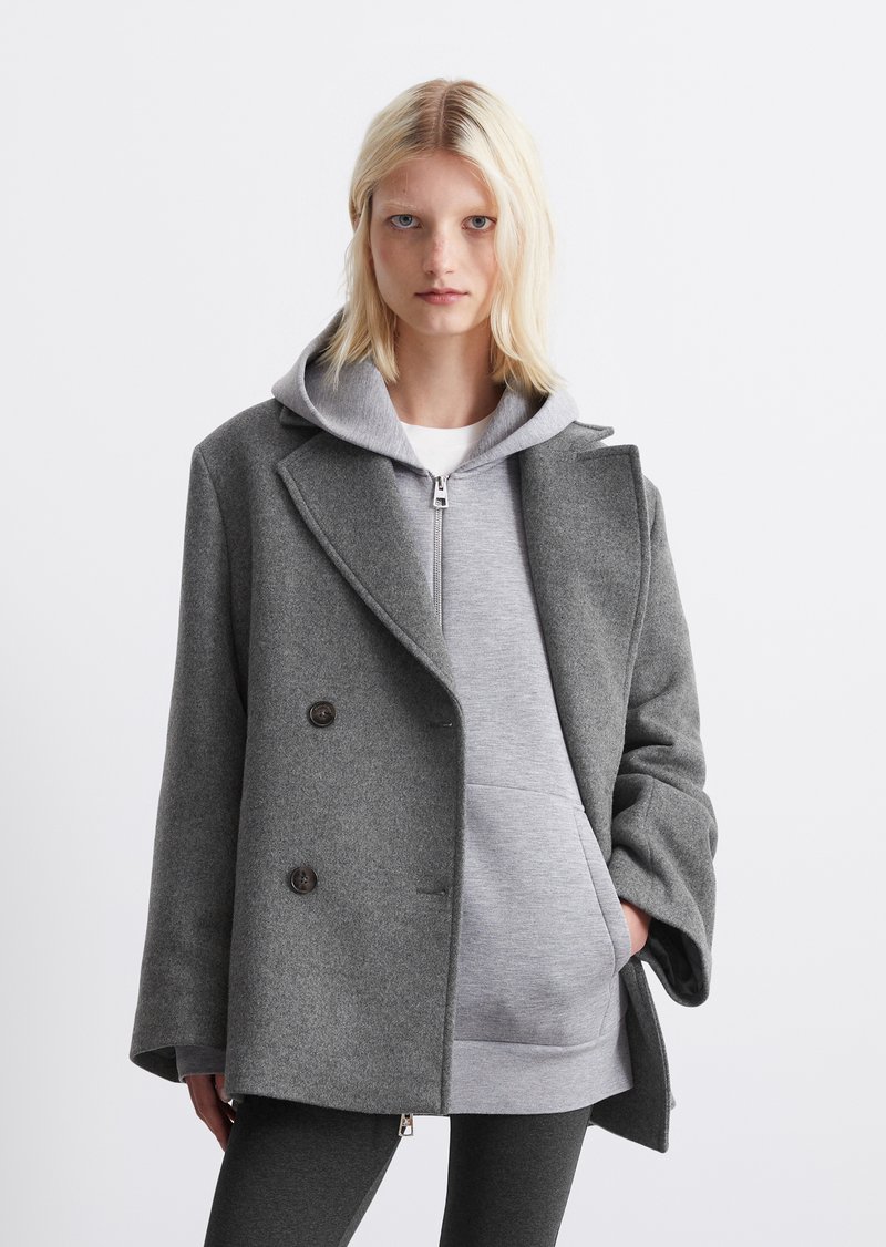 Cappotto grigio in lana doppiopetto con grandi bottoni, indossato sopra un hoodie grigio. Presenta una vestibilità comoda e un design con collare a punta.