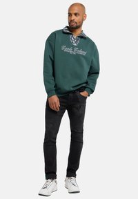 Groene trui met een patroon op de kraag, met witte tekst. Gecombineerd met zwarte jeans en witte schoenen, wat een casual stijl uitstraalt.