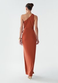 Asymetrické šaty na jedno rameno v cihlové barvě, dlouhé až na zem, s přiléhavým střihem, bočním vázáním a hladkým povrchem.