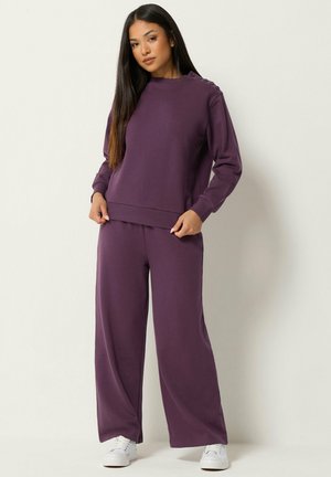 Vrouw staand tegen een effen achtergrond, gekleed in een losse paarse trui met knopen op één schouder en bijpassende wijde broek met witte sneakers.