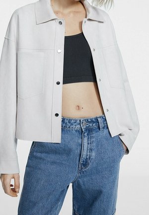 Vrouw draagt een lichtgrijze jas met knopen over een zwart cropped topje en hoog getailleerde blauwe spijkerbroek met één hand in de zak.