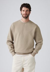 Beige sweatshirt met geribde kraag en boorden, met een subtiele logo op de borst. Draagt het met lichtgekleurde broeken, wat een casual uitstraling geeft.