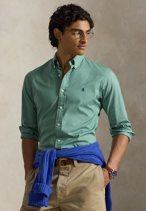 Uomo che indossa una camicia verde con bottoni e maniche arrotolate, pantaloni beige, maglione blu annodato in vita, occhiali tondi e un orologio da polso.