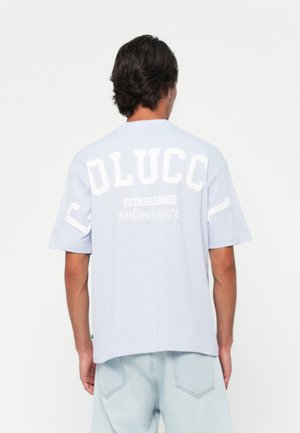 Mann iført en lyseblå t-skjorte med "OLUCC," "ETABLERT," og "Milano 1978" trykt i hvitt på ryggen, kombinert med lyseblå jeans.