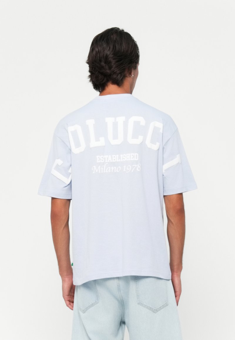 Mann iført en lyseblå t-skjorte med "OLUCC," "ETABLERT," og "Milano 1978" trykt i hvitt på ryggen, kombinert med lyseblå jeans.