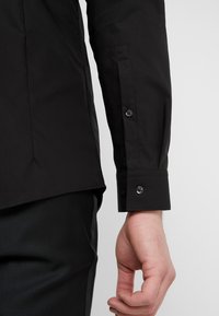 Zwart shirt met lange mouwen, voorzien van een getailleerd ontwerp, knoopmanchetten en een zachte textuur. Zichtbare stiksels aan de onderrand.