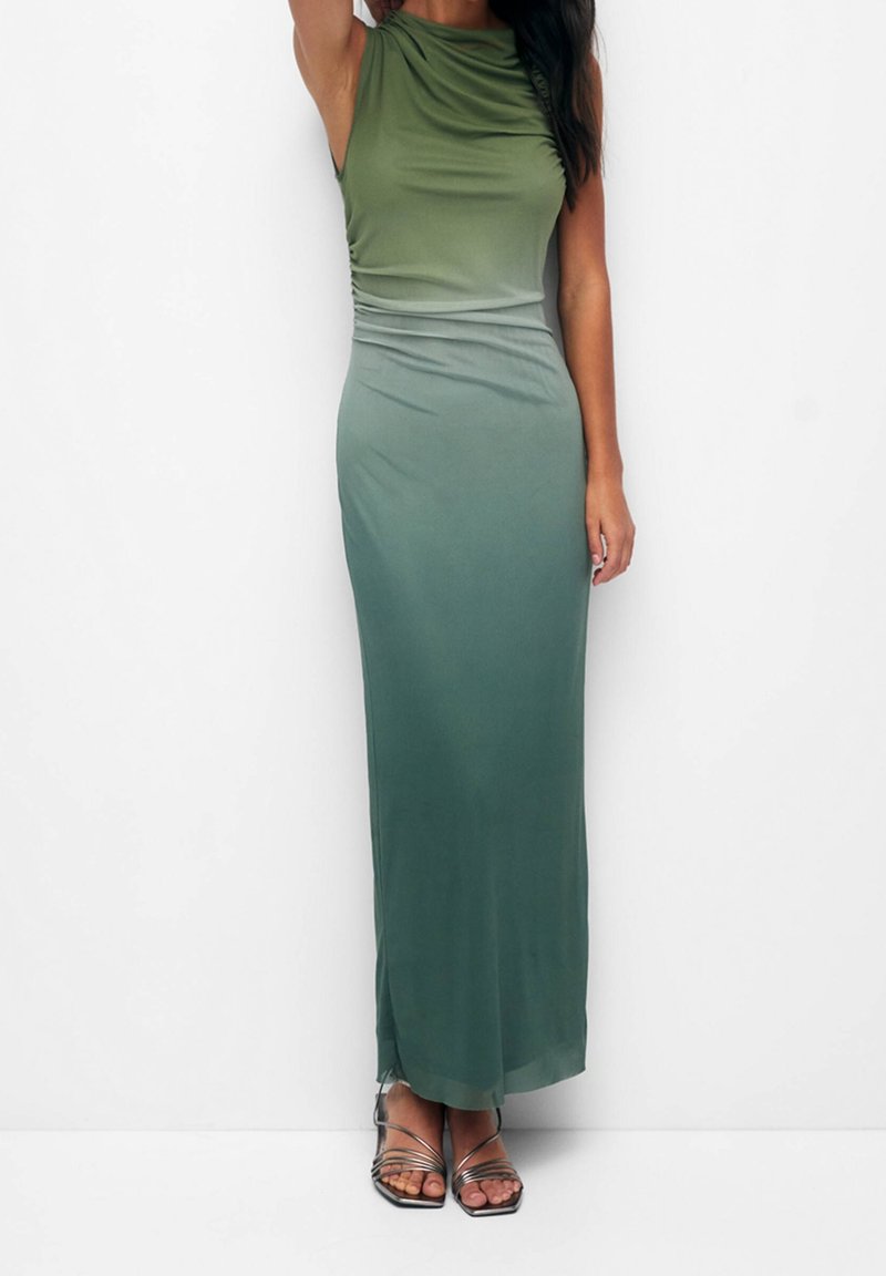 Grünes Gradientenkleid mit ärmellosem Design. Weicher, dehnbarer Stoff fällt sanft und zeigt eine geraffte Textur an der Seite. Silbergraue Sandalen mit Riemchen.