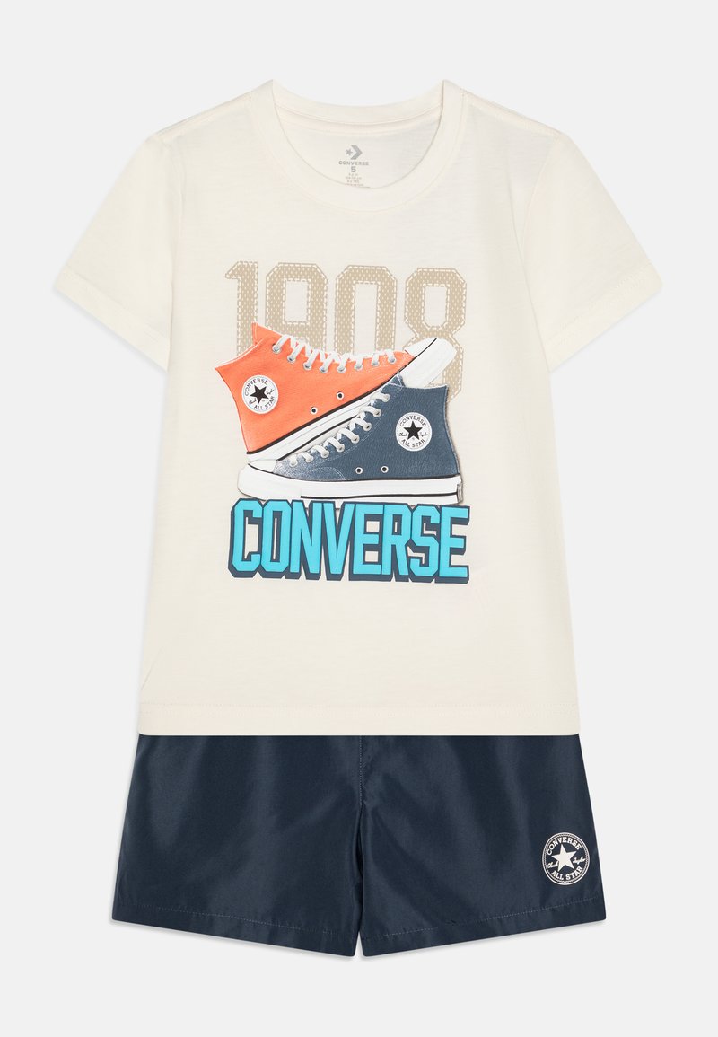 T-shirt en coton crème avec un graphique de baskets orange et marine, texte "1908" au-dessus, associé à un short marine avec un patch logo.