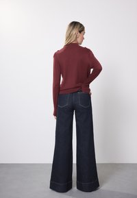 Pruunikas-tumesinine pikkade varrukatega kampsun, millel on liibuvad õlad, kombineeritud laiade säärega tumesiniste teksapükstega. Sile tekstuur, kontrastõmblused ja tagataskud.