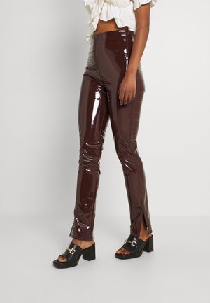 Kobieta ubrana w wysokie, błyszczące, brązowe spodnie winylowe z przednimi rozcięciami oraz czarne sandały na obcasie z odkrytymi palcami, stoi na tle białego tła.