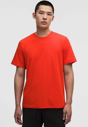 Giovane uomo che indossa una maglietta rossa a maniche corte e tinta unita con pantaloni neri, in piedi davanti a uno sfondo grigio chiaro uniforme.