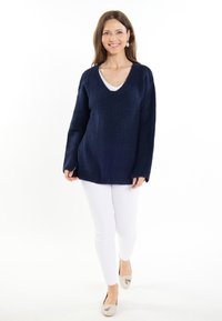 Tengerész kék, V nyakú, hosszú ujjú kötött pulóver, lazább szabással, fehér, slim-fit nadrággal és világos színű lapos cipőkkel párosítva.