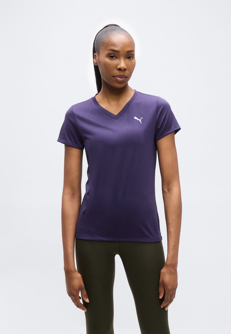 Γυναίκα που στέκεται φορώντας μοβ κοντομάνικη μπλούζα Puma και σκούρο πράσινο κολάν σε απλό λευκό φόντο.