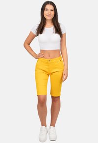 Geel denim shorts met een aangesloten ontwerp, voorzien van een knoopsluiting en vijf zakken, gedragen met een witte crop top en witte sneakers.