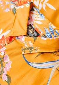 Chemise à manches courtes en tissu orange vif avec des motifs floraux rose et bleu, dotée d'un col et d'accents en boutons blancs.
