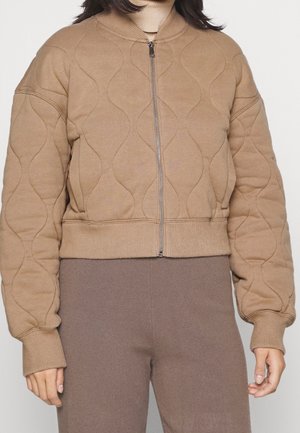 Person trägt eine beige gesteppte Jacke mit Reißverschluss, gerippten Bündchen und Saum, kombiniert mit braunen, hoch taillierten Hosen.