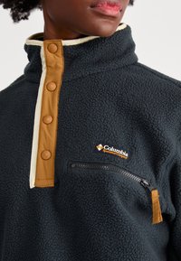 Pull en polaire navy foncé avec un col haut, une patte de boutonnage beige et une petite poche poitrine zippée. Logo Columbia affiché de manière proéminente.