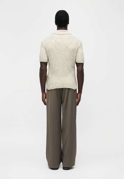 Άνδρας που φοράει υφασμάτινο μπεζ πουλόβερ με κοντά μανίκια και καφέ παντελόνι φαρδιάς γραμμής, στέκεται με την πλάτη προς τα εμπρός σε απλό φόντο.