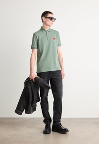 Lys grønn polo skjorte med tre knapper og rød etikett, sorte jeans, holder en svart denimjakke, og har på seg sorte solbriller og sko.