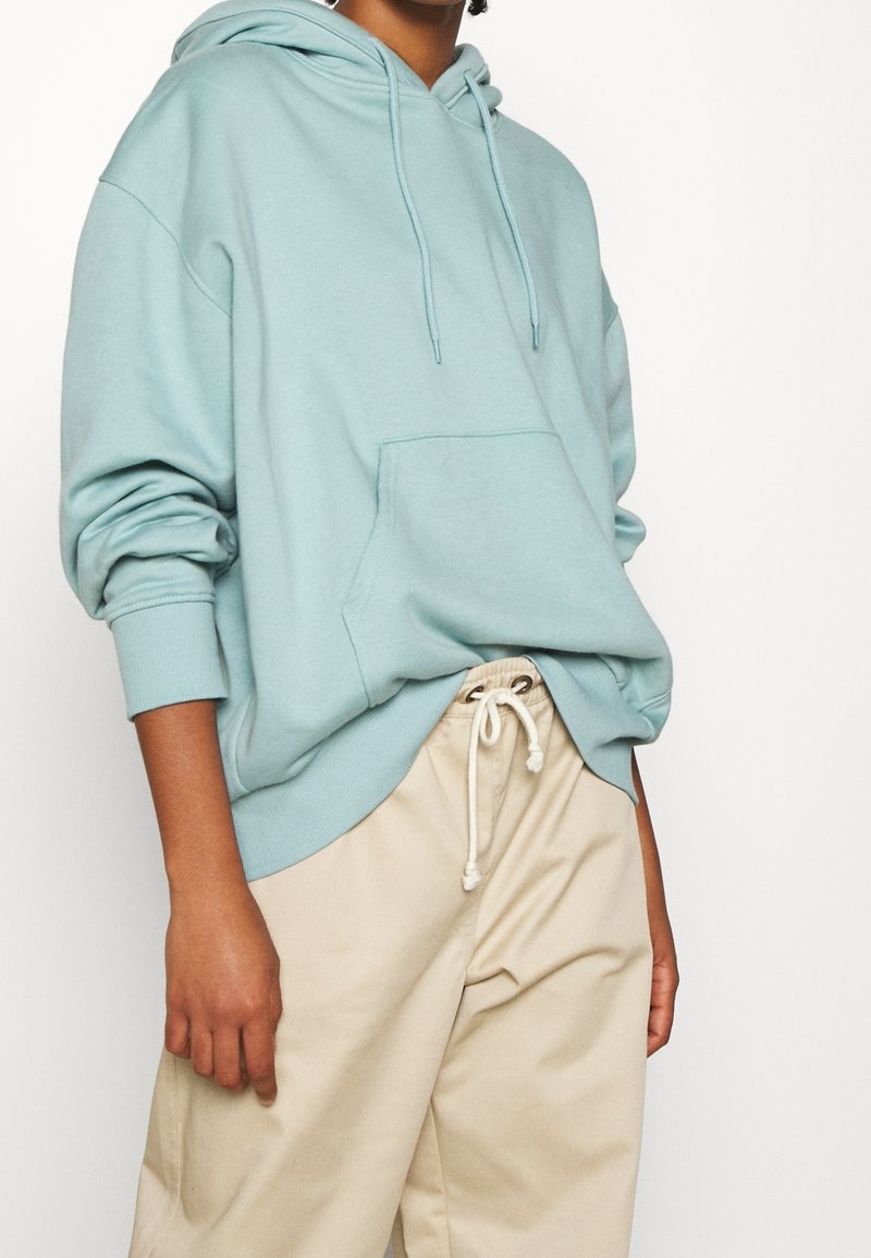 Persona que lleva una sudadera oversized de color azul claro con bolsillo frontal y pantalones de drawstring beige, de pie contra un fondo blanco.