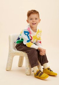 Maglione lavorato a maglia con motivi colorati, base bianca; pantaloni in velluto a coste marroni; scarpe gialle con strappi in Velcro; sedia maculata. Bambino seduto, sorridente.