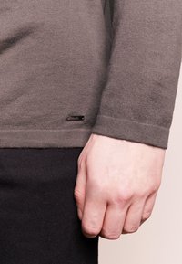 Maglione a maniche lunghe, lavorato a maglia marrone con orlo a coste, mostrato accanto a una mano. Presenta un piccolo cartellino nero del marchio vicino al fondo.