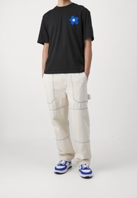 Camiseta de algodón negra con un gráfico de una flor sonriente azul, combinada con pantalones beige de corte holgado que presentan costuras azules y bolsillos laterales.